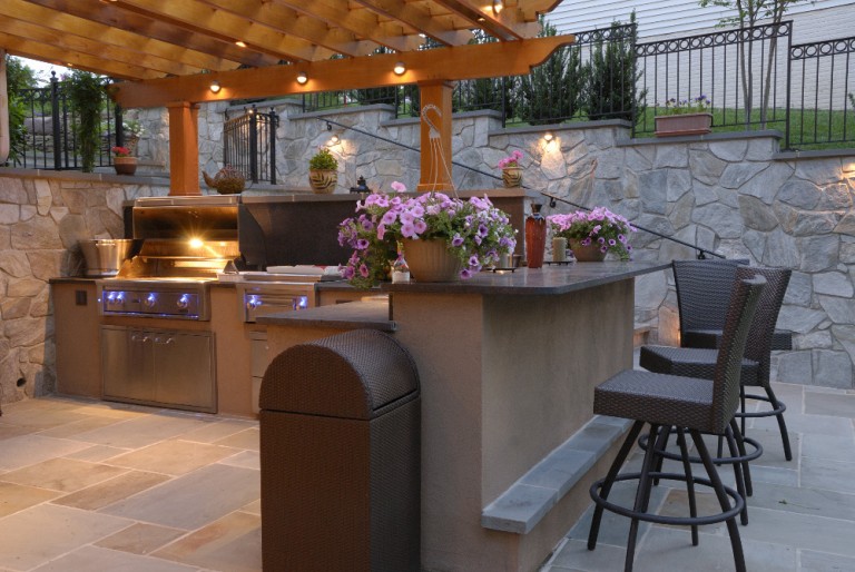 Outdoor kitchen with grill and bar seating. Stone pavers and wood pergola.
