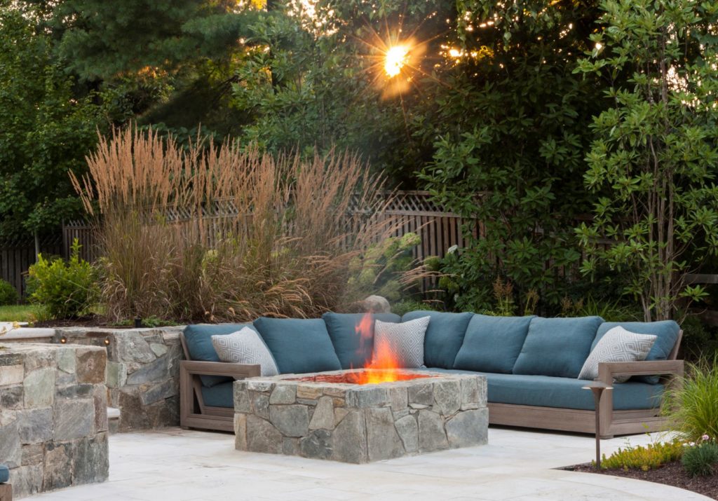 Custom-built backyard patio and fire pit with blue patio sectional next to it, and trees and setting sun in background.