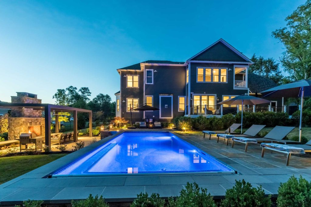 Large inground swimming pool installed behind large two-story house at dusk. Lights on inside house.