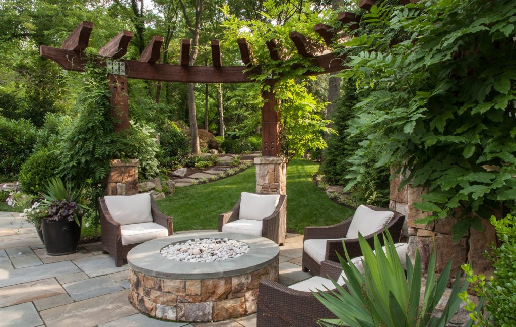 Outdoor patio with built-in stone fire pit surrounded by cushioned patio chairs and lots of trees and greenery around.