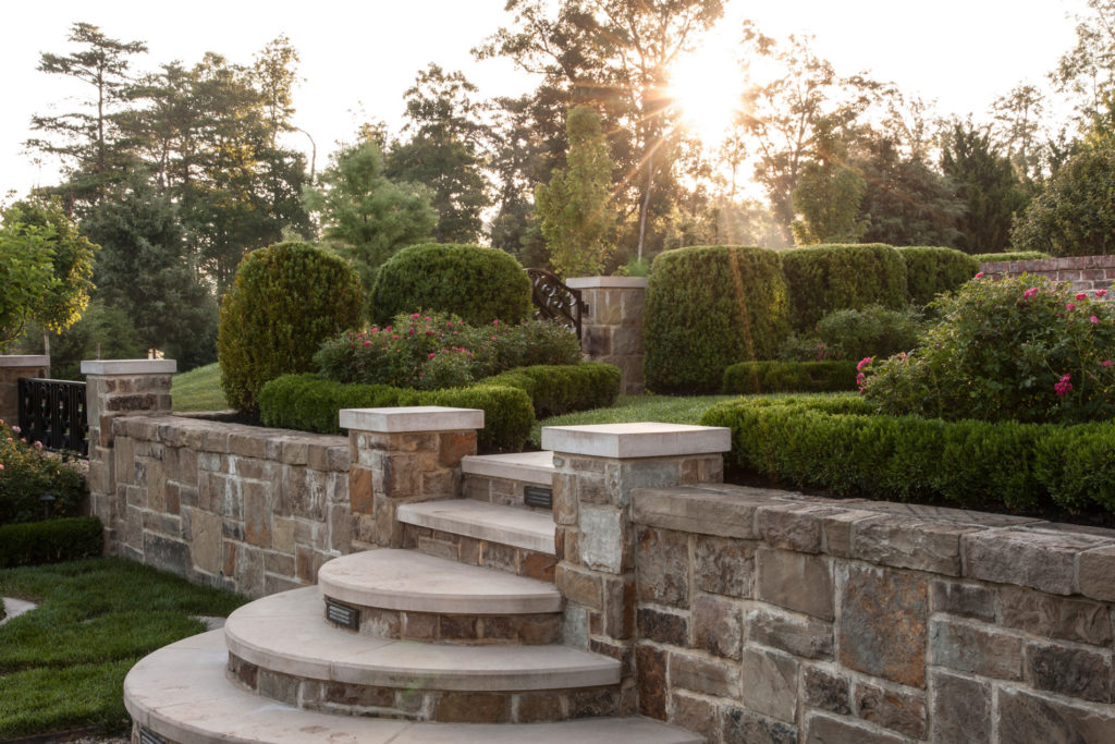 Stone retaining wall with set of rounded stone steps leading up to manicured garden past retaining wall. Sunset.