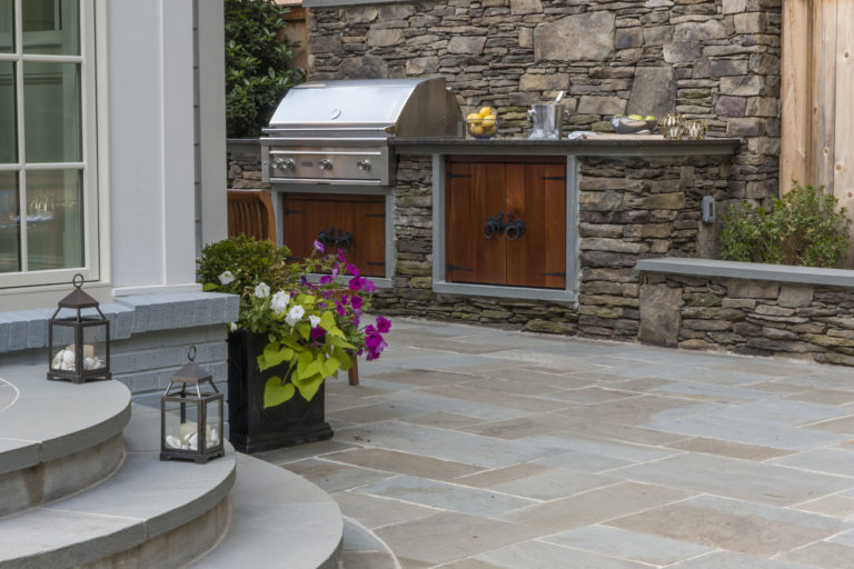 Outdoor kitchen with covered grill, wood-grain cabinets set in stone wall along side of stone patio.