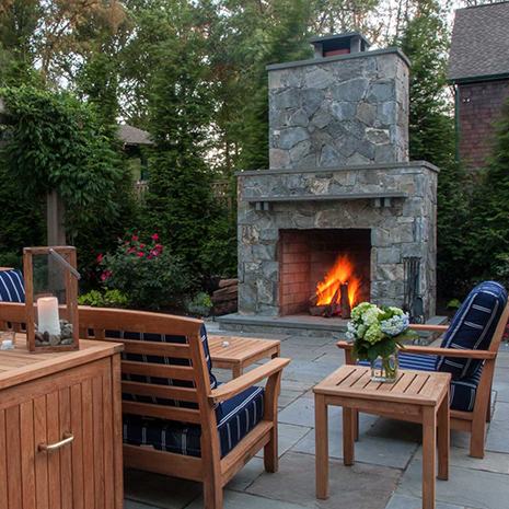 Wooden furniture and a fire place on a patio, set against a backdrop of lush landscape and flowers.
