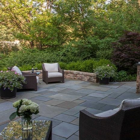 Scenic patio featuring wicker furniture, fire pit, and vibrant potted plants amidst lush landscape.