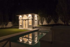 Landscaped yard with inground pool and small pool house lit up at night.