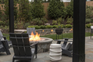 Gray Adirondack chairs around stone firepit in landscaped yard. 