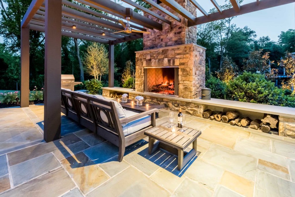 Outdoor patio with large stone fireplace set into low stone wall, with patio sofa facing it.
