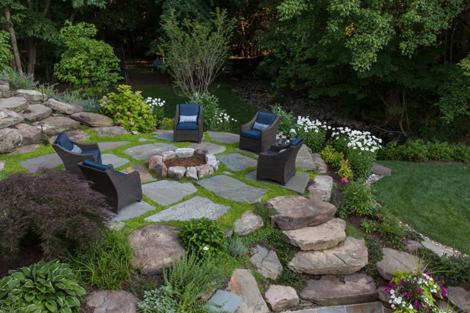 Large backyard with stone walkway and chairs around a stone firepit. 