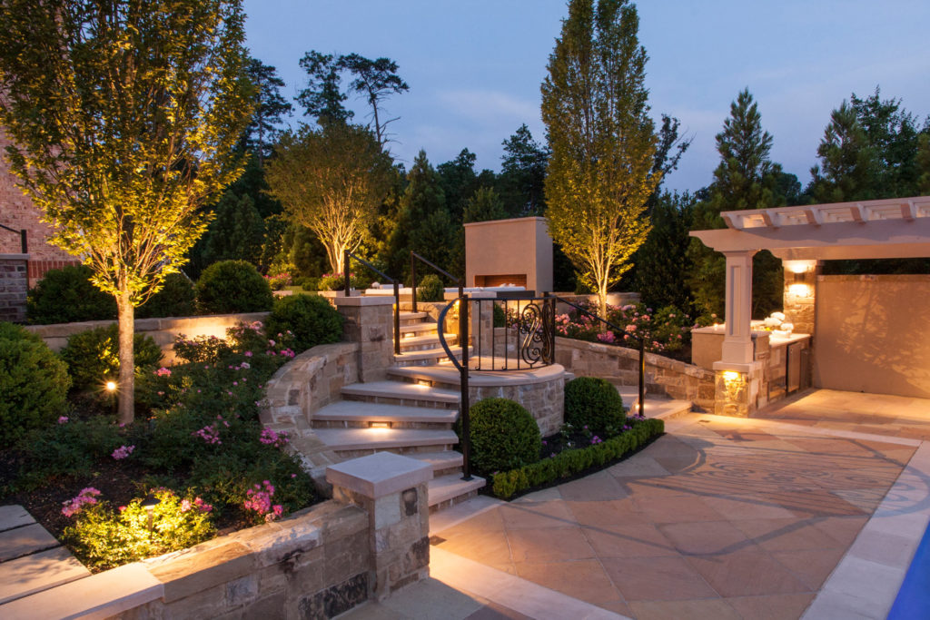 A stone walkway with a staircase and gate with custom outdoor lighting on the stairs and surrounding garden beds
