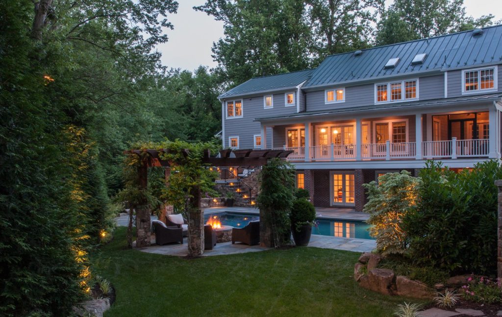 A serene backyard patio at dusk, with a stone wall and stone bench, a small pergola, a large in-ground pool, all behind a three-story home with large back patio and lights on inside.