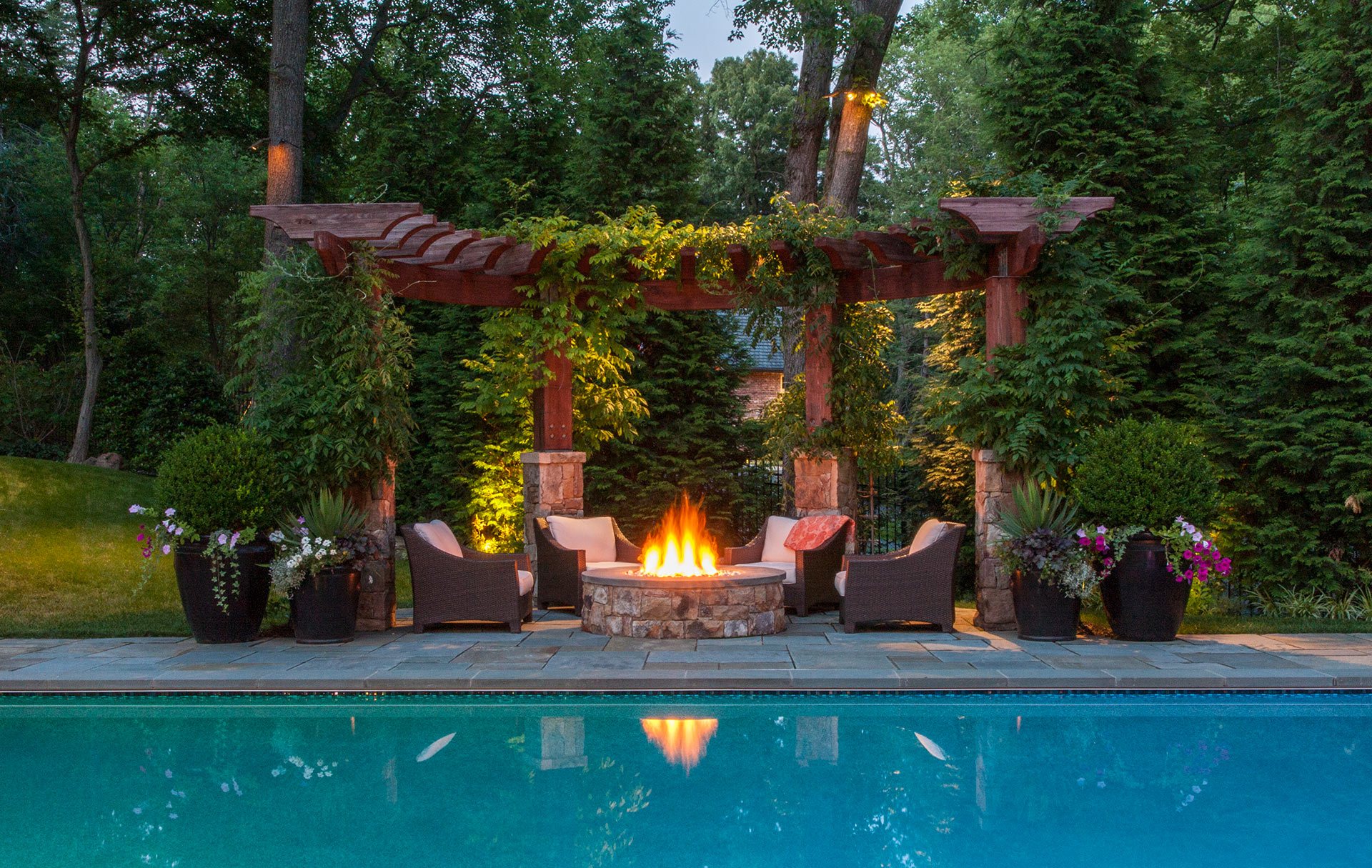Pool and a gazebo with a fireplace surrounded by lush trees.