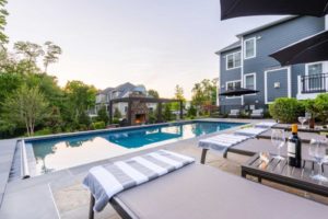 View of large inground pool and patio with lounge chairs behind large blue house.