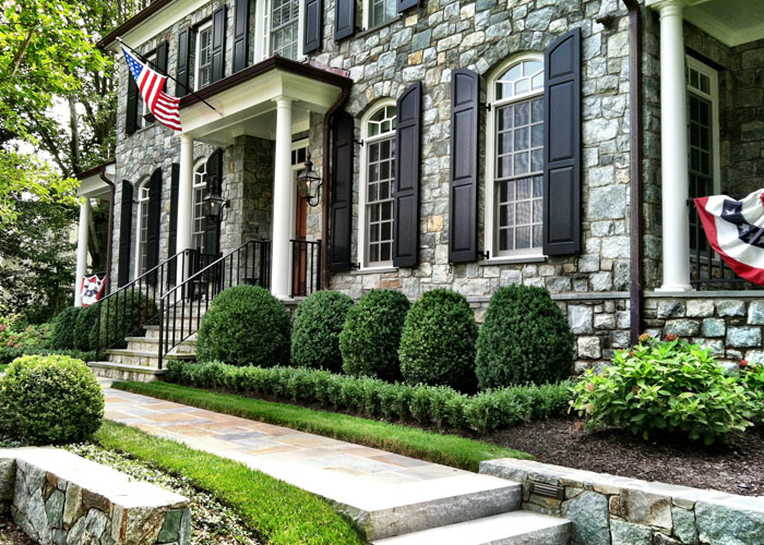 Landscaping, walkway design leading up to large stone home with black shutters and landscaped shrubbery in front.